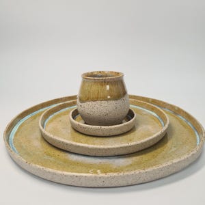 Könnte beinhalten: Ein Keramik-Geschirrset mit einer gesprenkelten, hellbraunen Tasse und passenden Tellern. Die Teller haben einen hellblauen Rand und eine gesprenkelte, hellbraune Oberfläche. Die Tasse ist zweifarbig, mit einem hellbraunen Oberteil und einem gesprenkelten, cremefarbenen Boden.