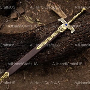 May include: An ornate sword with a gold-colored hilt and blade guard, featuring intricate designs and a blue gem. The blade is housed in a brown sheath with gold accents. The sword rests on a dark, textured wooden surface.