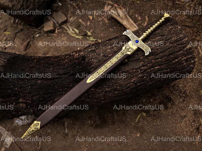 May include: An ornate sword with a gold-colored hilt and blade guard, featuring intricate designs and a blue gem. The blade is housed in a brown sheath with gold accents. The sword rests on a dark, textured wooden surface.