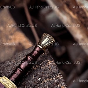 May include: A close-up of a decorative sword with a gold-colored hilt and accents. The handle is wrapped in a dark burgundy leather. The blade is partially visible, with gold-colored detailing and text. The sword rests on a weathered wooden surface.