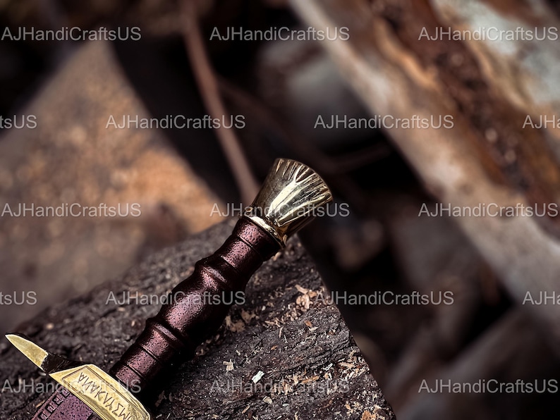 May include: A close-up of a decorative sword with a gold-colored hilt and accents. The handle is wrapped in a dark burgundy leather. The blade is partially visible, with gold-colored detailing and text. The sword rests on a weathered wooden surface.