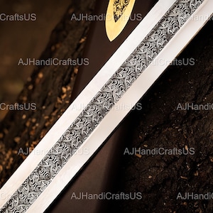 May include: Close-up of a sword blade with a detailed, ornate design running down the center. The blade is silver, and the central design is black and silver. The text "AJHandiCraftsUS" is visible in the image.