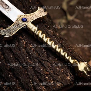 May include: A decorative sword with a gold-colored handle and crossguard, featuring intricate engravings and a blue gem. The blade is silver and the sword rests on a dark, textured surface.
