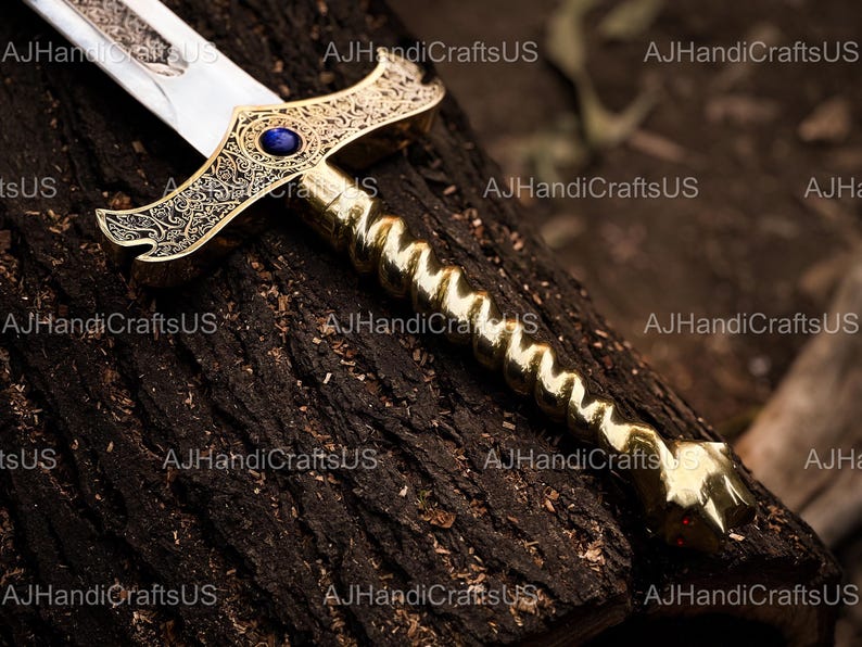 May include: A decorative sword with a gold-colored handle and crossguard, featuring intricate engravings and a blue gem. The blade is silver and the sword rests on a dark, textured surface.