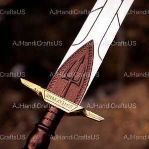 May include: A close-up of a decorative sword with a bronze handle and a silver blade. The sword features a bronze guard with the Greek word "ANAKLYSMOS" and a trident symbol. The blade is white with etched lines.