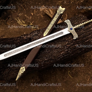 May include: A decorative sword with a silver blade and gold accents, resting on a dark brown surface. The hilt features a blue gem. The sword is accompanied by a brown sheath with gold detailing. The text "AJHandiCraftsUS" is visible.
