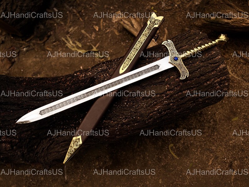 May include: A decorative sword with a silver blade and gold accents, resting on a dark brown surface. The hilt features a blue gem. The sword is accompanied by a brown sheath with gold detailing. The text "AJHandiCraftsUS" is visible.