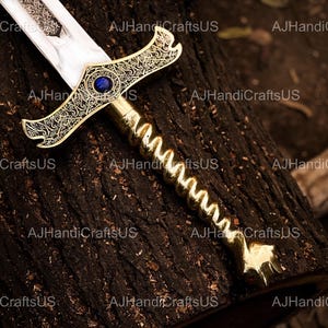 May include: A decorative sword with a gold-colored handle and crossguard, featuring intricate scrollwork and a blue gem. The blade is silver and etched. The sword rests on a dark brown wooden surface.