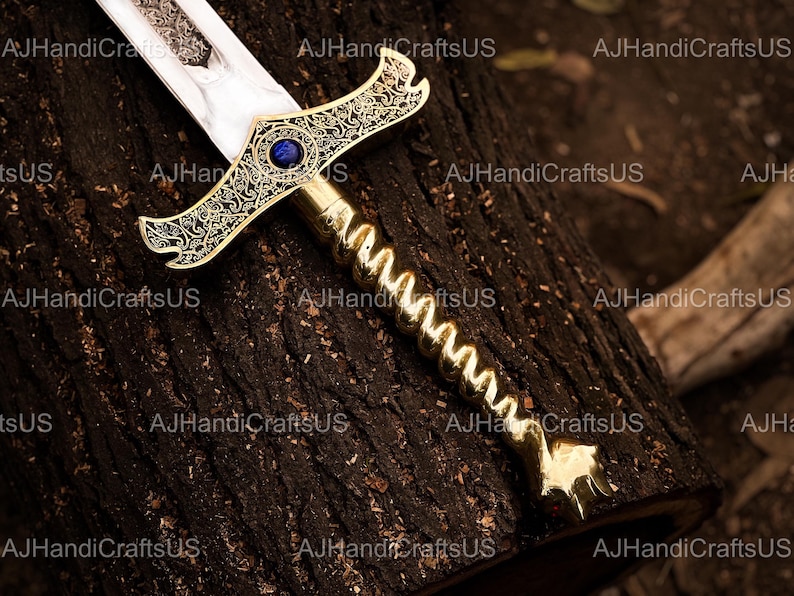 May include: A decorative sword with a gold-colored handle and crossguard, featuring intricate scrollwork and a blue gem. The blade is silver and etched. The sword rests on a dark brown wooden surface.
