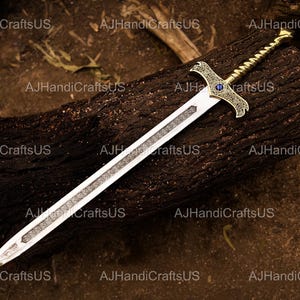 May include: A decorative sword with a silver blade and ornate gold hilt. The hilt features a blue gem and intricate detailing. The sword rests on a dark brown, textured surface. The text "AJHandiCraftsUS" is visible.