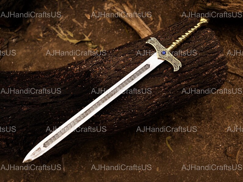 May include: A decorative sword with a silver blade and ornate gold hilt. The hilt features a blue gem and intricate detailing. The sword rests on a dark brown, textured surface. The text "AJHandiCraftsUS" is visible.