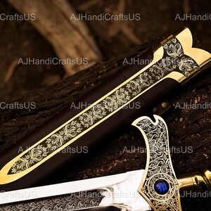 May include: A decorative sword with a gold-colored hilt and blade, featuring intricate floral patterns. The sword's handle has a blue gem. The scabbard is dark brown. The sword is displayed on a dark, textured surface.