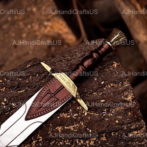 May include: A detailed image of a decorative sword with a brown leather-wrapped handle and a gold-colored hilt. The blade is silver with a brown leather guard featuring a trident design. The sword rests on a textured wooden surface.