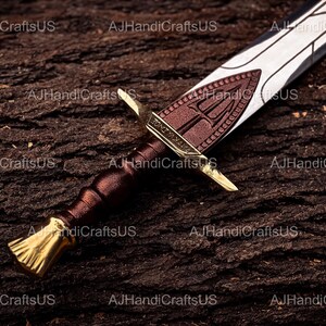 May include: A close-up of a decorative sword with a brown handle and a gold-colored hilt. The blade is silver with a brown emblem. The sword rests on a textured, dark brown surface.