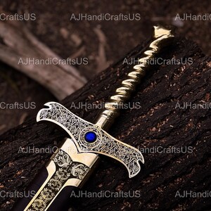 May include: A decorative sword with a gold-colored hilt and blade, featuring intricate engraved patterns and a blue gem. The sword rests on a dark, textured wooden surface, showcasing its ornate design.