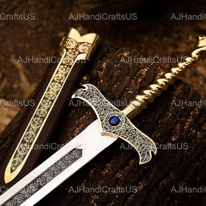 May include: An ornate sword with a gold handle and a silver blade rests on a wooden surface. The hilt features intricate designs and a blue gem. The scabbard is brown with gold accents. The sword is a decorative item.