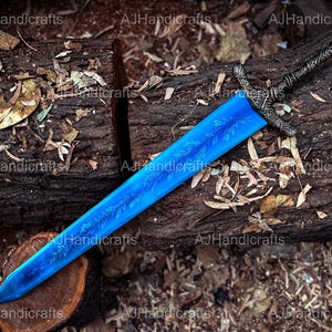 May include: A blue sword with a silver handle and intricate floral designs etched into the blade. The sword is lying on a wooden surface with leaves and twigs scattered around it.