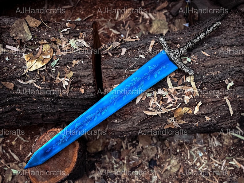 May include: A blue sword with a silver handle and intricate floral designs etched into the blade. The sword is lying on a wooden surface with leaves and twigs scattered around it.