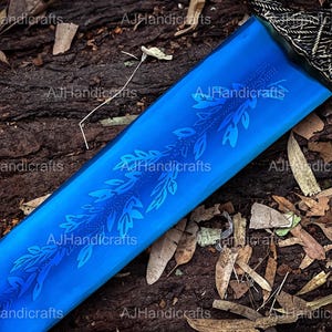 May include: A close-up of a blue sword blade with a floral design etched into the surface. The blade is resting on a wooden surface with brown leaves scattered around it.