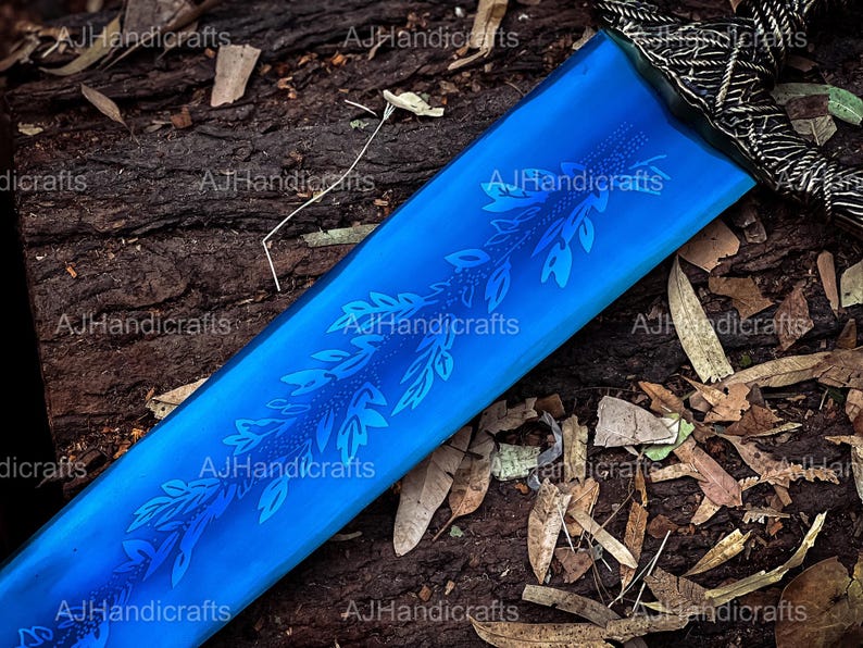 May include: A close-up of a blue sword blade with a floral design etched into the surface. The blade is resting on a wooden surface with brown leaves scattered around it.