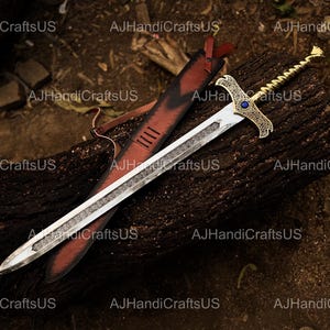 May include: A decorative sword with a silver blade and ornate gold hilt, featuring a blue gem. The sword rests on a dark wooden surface next to a red leather sheath. The blade has a detailed etched design.