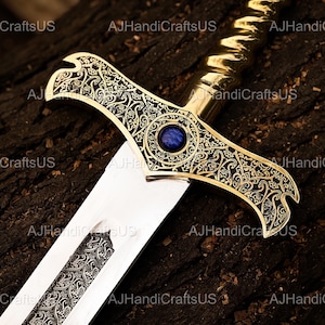 May include: A decorative sword with a gold-colored hilt and crossguard, featuring intricate engraved patterns. The crossguard has a blue gemstone in the center. The blade is silver with engraved details. The sword rests on a dark, textured surface.