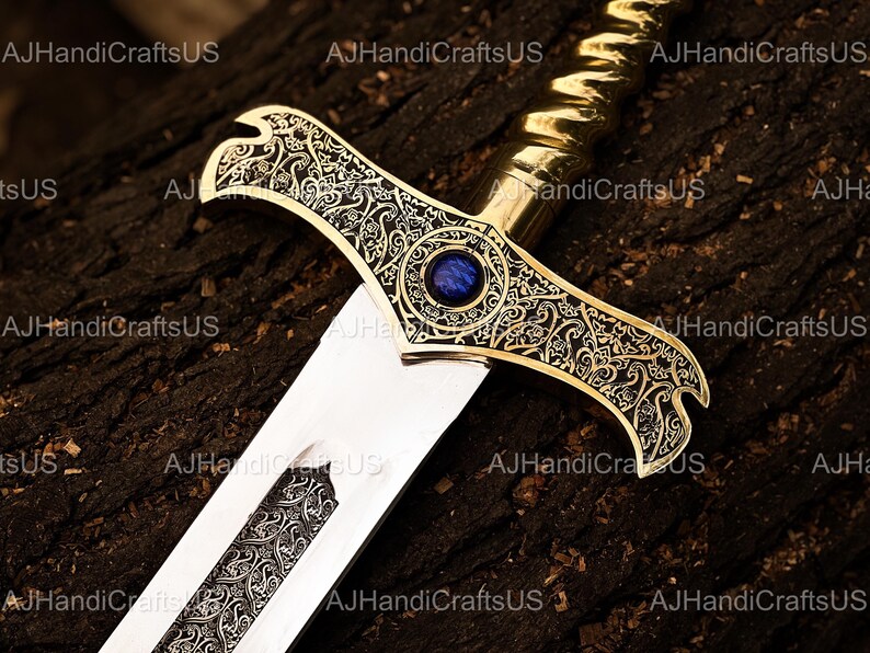 May include: A decorative sword with a gold-colored hilt and crossguard, featuring intricate engraved patterns. The crossguard has a blue gemstone in the center. The blade is silver with engraved details. The sword rests on a dark, textured surface.
