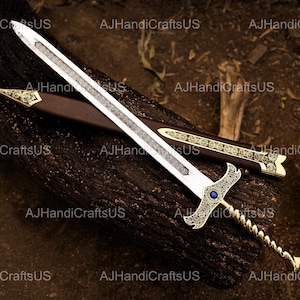 May include: A decorative sword with a silver blade and ornate gold handle, resting on a dark brown wooden surface. The sword features a blue gem and is accompanied by a brown sheath with gold accents. The text "AJHandiCraftsUS" is visible.