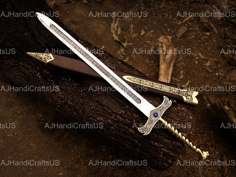May include: A decorative sword with a silver blade and ornate gold handle, resting on a dark brown wooden surface. The sword features a blue gem and is accompanied by a brown sheath with gold accents. The text "AJHandiCraftsUS" is visible.