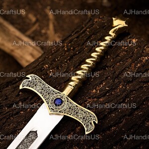 May include: A decorative sword with a gold-colored handle and crossguard, featuring intricate patterns and a blue gem in the center. The blade is silver and the sword rests on a dark, textured surface. The text "AJHandiCraftsUS" is visible.