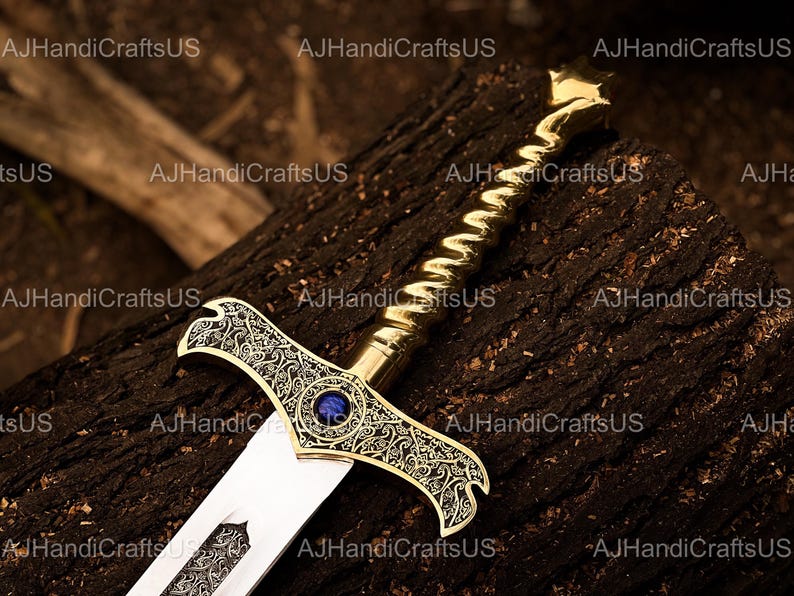 May include: A decorative sword with a gold-colored handle and crossguard, featuring intricate patterns and a blue gem in the center. The blade is silver and the sword rests on a dark, textured surface. The text "AJHandiCraftsUS" is visible.