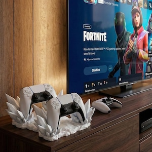 May include: A modern entertainment setup featuring a large screen displaying the Fortnite game interface. Two white and black game controllers rest on a decorative stand, with another controller nearby. The setup is on a dark wood cabinet.