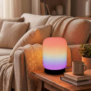 Op de afbeelding: Een cilindervormige lamp met een kleurengradiënt, van oranje naar paars, staat op een houten tafel. De lamp heeft een zwarte basis en is aangesloten. Een mok en boeken staan ook op de tafel.