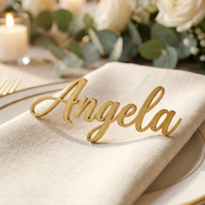 Pode incluir: Um marcador de lugar dourado, com o nome "Angela" em itálico, repousa sobre um guardanapo de linho branco sujo dobrado. O marcador de lugar está sobre um prato branco com borda dourada. O fundo inclui velas, flores e um castiçal dourado.