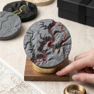 Könnte beinhalten: Eine graue topografische Karten-Skulptur mit roten Linien, montiert auf einem Holzsockel mit einem Messingring. Weitere Karten-Skulpturen in Schwarz, Grau und Beige sind sichtbar. Eine schwarze Geschenkbox und ein Kompass sind ebenfalls im Bild.