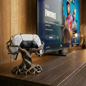 Könnte beinhalten: Ein weiß-schwarzer Gamecontroller liegt auf einem bronzefarbenen, baumförmigen Ständer. Der Ständer steht auf einer dunkelbraunen Holzoberfläche, im Hintergrund ist ein Fernseher mit dem Spiel "Fortnite" zu sehen. Eine Lampe ist ebenfalls sichtbar.