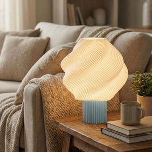 Può includere: Una moderna lampada da tavolo con un paralume a spirale color crema e una base blu chiaro a coste. La lampada è posizionata su un tavolo di legno accanto a una tazza e dei libri. Il design della lampada aggiunge un tocco contemporaneo alla stanza.