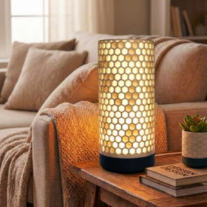Op de afbeelding: Een cilindrische tafellamp met een honingraatpatroon, die een warme gloed uitstraalt. De lamp staat op een donkere basis en staat op een houten tafel. Een kleine vetplant in pot en boeken staan ook op de tafel.