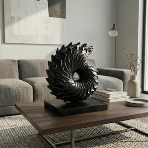 Könnte beinhalten: Eine dunkle, abstrakte Skulptur, die einer Nautilusmuschel ähnelt, steht auf einem quadratischen Sockel auf einem Holztisch. Die Skulptur ist schwarz und hat eine strukturierte Oberfläche. Das Wohnzimmer hat ein graues Sofa und eine neutrale Farbpalette.