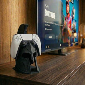 May include: A black and white PlayStation 5 controller resting on a black Spartan helmet-shaped stand. The controller is white with black buttons. The stand is on a dark brown wooden surface, with a TV in the background.