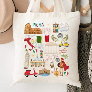 May include: A white canvas tote bag with a colorful design featuring Italian landmarks, including the Colosseum, the Trevi Fountain, the Leaning Tower of Pisa, and the Pantheon. The bag also features the words 'Roma', 'Grazie', 'La Dolce Vita', and 'Buongiorno'.