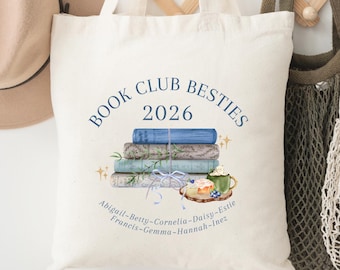 May include: A cream-colored canvas tote bag featuring a printed illustration of a stack of books, a mug, and a pastry. The text reads "BOOK CLUB BESTIES 2026" above the books, with a list of names including Abigail, Betty, Cornelia, and others below.