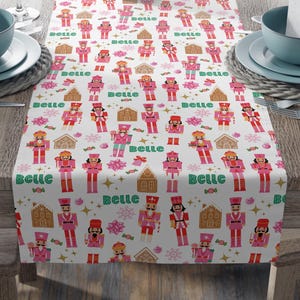 May include: A white table runner with a repeating pattern of pink nutcrackers, gingerbread houses, and snowflakes. The word "Belle" is written in green script throughout the design. The runner is set on a wooden table with place settings.