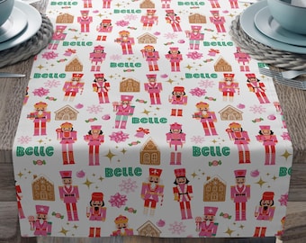 May include: A white table runner with a repeating pattern of pink nutcrackers, gingerbread houses, and snowflakes. The word "Belle" is written in green script throughout the design. The runner is set on a wooden table with place settings.