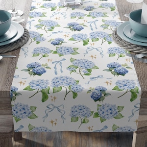 Könnte beinhalten: Ein weißer Tischläufer mit einem Aquarellmuster aus blauen Hortensien, Schleifen und goldenen Sternen. Der Läufer liegt auf einem Holztisch mit Gedecken, Weingläsern und einer Vase mit rosa Lilien. Das Gesamtthema ist floral und dekorativ.
