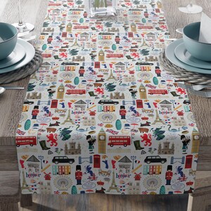 May include: A cream-colored table runner with a repeating pattern of London and Paris landmarks, including the Eiffel Tower, Big Ben, and red double-decker buses. The runner is set on a wooden table with place settings and a vase of pink flowers.