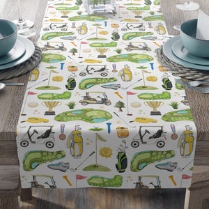Puede incluir: Camino de mesa blanco con un diseño temático de golf. El diseño incluye carritos de golf, bolsas de golf, banderas y elementos del campo de golf en verde, amarillo y marrón. El camino de mesa está sobre una mesa de madera.