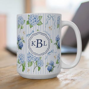 May include: White ceramic mug with a blue and white striped pattern, decorated with watercolour blue hydrangea flowers, bows, and stars. The mug features a circular emblem with the initials "KBL".