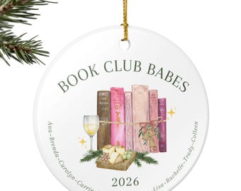 Peut inclure: Une décoration ronde en céramique blanche avec le texte BOOK CLUB BABES et 2026. Le motif montre une rangée de livres colorés, un verre de vin blanc et un plateau de fromages. Une liste de noms entoure le bas et un cordon doré est fixé.