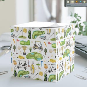May include: A white cube-shaped notepad with a golf-themed pattern. The design includes golf carts, golf balls, golf clubs, and green golf course illustrations. The notepad is on a white desk with office supplies.